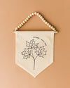 A beaded banner with a black and white illustration of three maple leaves hanging on a wall.