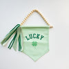Green banner with 'LUCKY' and clover leaf design, hanging from wooden beads on a white background
