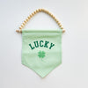 Green banner with 'LUCKY' and clover leaf design, hanging from wooden beads on a white background